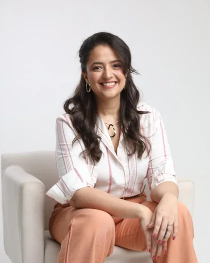 Dra. Kamila Giraldelli Junqueira sorrindo, de camisa branca listrada, sentada numa poltrona clara em estúdio de fundo cinza claro